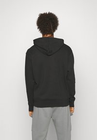 Czarna bluza z kapturem ze sznurkiem i ściągaczami na mankietach, wykonana z miękkiego materiału. Pokazana od tyłu, o prostym i czystym wzornictwie.