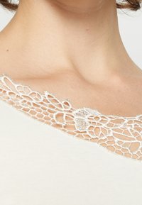 Weißes Oberteil mit aufwändigen Spitze-Details am Ausschnitt, einem zarten Blumenmuster und weicher Textur.