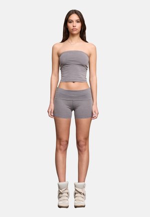 Femme debout face à l’objectif, portant un haut bustier gris, un short assorti et des baskets montantes beige clair sur fond uni.
