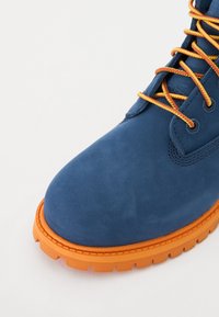 Sinine nahast ankle king, millel on tekstuuriga pind, oranž kummitald ja kontrastselt oranžid paelad. Profiilipilt toob esile ümarad varbad.