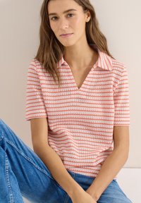 Jeune femme aux longs cheveux bruns portant un haut tricoté à manches courtes rayé corail et blanc, et un jean bleu, assise devant un fond clair.