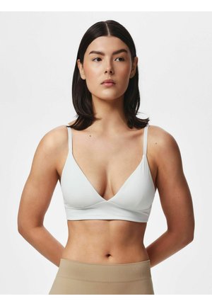 Žena s rovnými tmavými vlasmi, ktorá má na sebe biely bralette a béžové nohavice s vysokým pásom, stojí s rukami za chrbtom na jednofarebnom pozadí.
