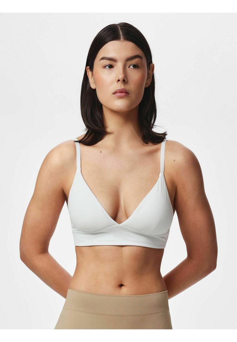 Femme aux cheveux lisses et foncés portant un bralette blanc et un pantalon beige taille haute, debout les mains derrière le dos sur un fond uni.