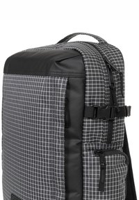 Zaino in tessuto a scacchi bianco e nero con parte superiore imbottita, zip laterali, spallacci regolabili e dettaglio con fibbia frontale.