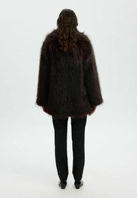 Abrigo de piel sintética en burdeos oscuro, diseño oversize con un acabado suave y texturizado. Combinado con pantalones y zapatos negros, visto desde la parte de atrás.
