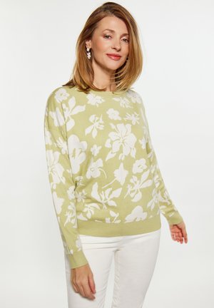Maglione verde chiaro con motivo floreale oversize in bianco, scollatura rotonda, spalle basse e polsini e orlo a coste. Abbinato a pantaloni bianchi.