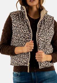 Chaleco de piel de leopardo con un cuello crema y cremallera, llevado sobre un suéter marrón texturizado. Presenta una forma ajustada y detalles acolchados.