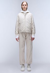 Lichtbeige ritsjack met capuchon, gecombineerd met bijpassende losvallende beige broek en witte schoenen. Gladde stof met subtiele details.