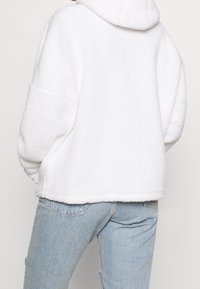 White, fluffy fleece hoodie with a hood, wide sleeves, and a relaxed fit. Paired with light blue, distressed denim jeans.