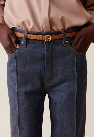 Jeans de mezclilla azul oscuro con costuras naranjas en contraste, que presentan un cinturón de cuero marrón suave con una hebilla dorada y bolsillos frontales.