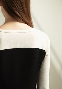 Femme aux longs cheveux bruns portant un haut texturé noir et blanc, vue de dos sur un fond clair neutre.