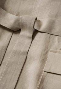 Vêtement en lin beige avec ceinture, surface texturée et poche latérale. Le design comprend des coutures visibles et une finition soignée.