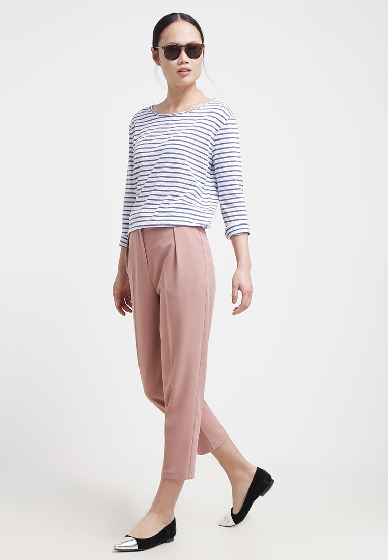 Frau trägt Sonnenbrille, gestreiftes Langarmshirt, rosa verkürzte Hose und schwarze Ballerinas mit silbernen Zehen, steht vor einem weißen Hintergrund.