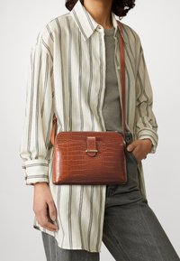 Brown faux-crocodile leather crossbody bag featuring a rectangular shape, zip closure, and adjustable strap. Worn with a striped shirt.