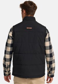 Chaleco tipo puffer negro con cuello alto, que presenta una etiqueta de cuero rectangular, usado sobre una camisa a cuadros en colores negro y crema.