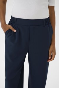 Persona valkā tumši zili elastīgus bikses ar sānu kabatām, roka kabatā, un baltu rībveida īsroku kreklu.