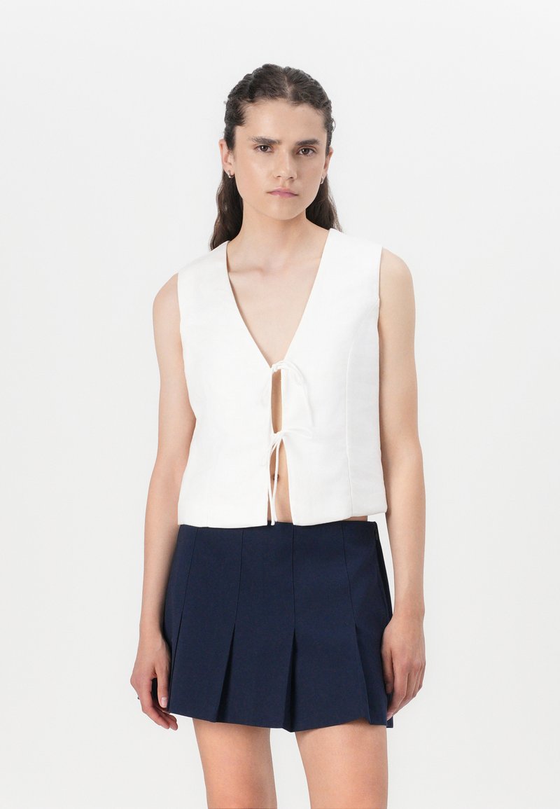Witte mouwloze cropped top met een diepe V-hals en striksluiting aan de voorkant, gecombineerd met een geplooide marineblauwe rok. Glad stoftextuur.