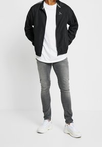 Svart zip-up jacka med hög krage, bärs över en vit t-shirt. Grå slim-fit jeans och vita sneakers kompletterar outfiten.