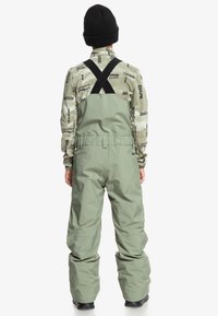 Groene waterdichte overall met een losse pasvorm, verstelbare zwarte bandjes, een gemêleerde grijze fleece eronder en een zwarte beanie op het hoofd.
