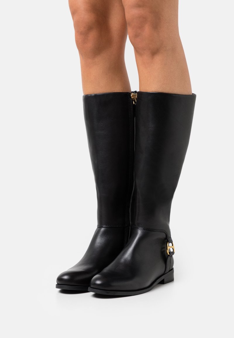 Bottes en cuir noires jusqu'aux genoux avec un détail de boucle dorée portées sur des jambes nues, montrées sur un fond clair uni.