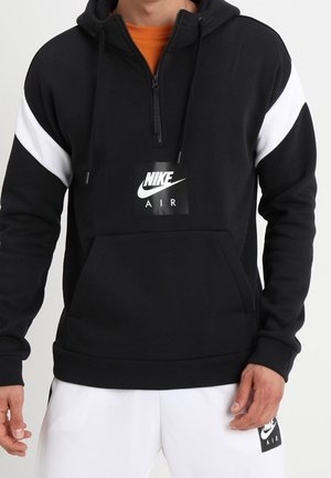 Homme portant un sweat à capuche Nike Air noir avec des bandes blanches sur les manches et un short Nike Air blanc, debout devant un fond uni.