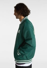 Vihreä pommikaukalo, jossa on raidalliset resorit hihansuissa ja kauluksessa. Varustettu edessä vetoketjulla, sivutaskuilla ja hienovaraisella brodeeratulla kuvioinnilla selkäpuolella.