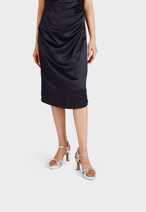 Donna che indossa un vestito nero in raso fino al ginocchio e sandali argentati con tacco alto e cinturini alla caviglia su uno sfondo neutro.