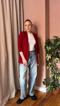 Γυναίκα με κόκκινο σακάκι, λευκό πουκάμισο, ανοιχτά μπλε τζιν και μαύρα παπούτσια, που στέκεται σε εσωτερικό χώρο δίπλα σε μπεζ κουρτίνες και μια γλάστρα με ποικιλόχρωμο φυτό.