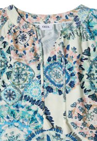 Blouse blanche avec des motifs floraux abstraits bleu, sarcelle et pêche, avec un col rond et deux liens à pompons suspendus à l'avant.