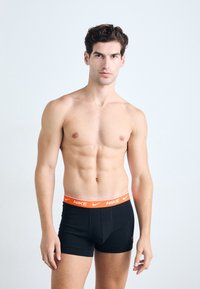 Fekete Nike boxeralsó narancsszínű derékpánttal, szoros illeszkedéssel és sima textúrával. A modell semleges arckifejezéssel pózol.