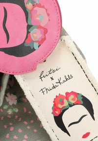 Pouch redondo rosa y artículo de tela con la cara estilizada de Frida Kahlo, flores y la firma "fritzi x Frida Kahlo" sobre lona.