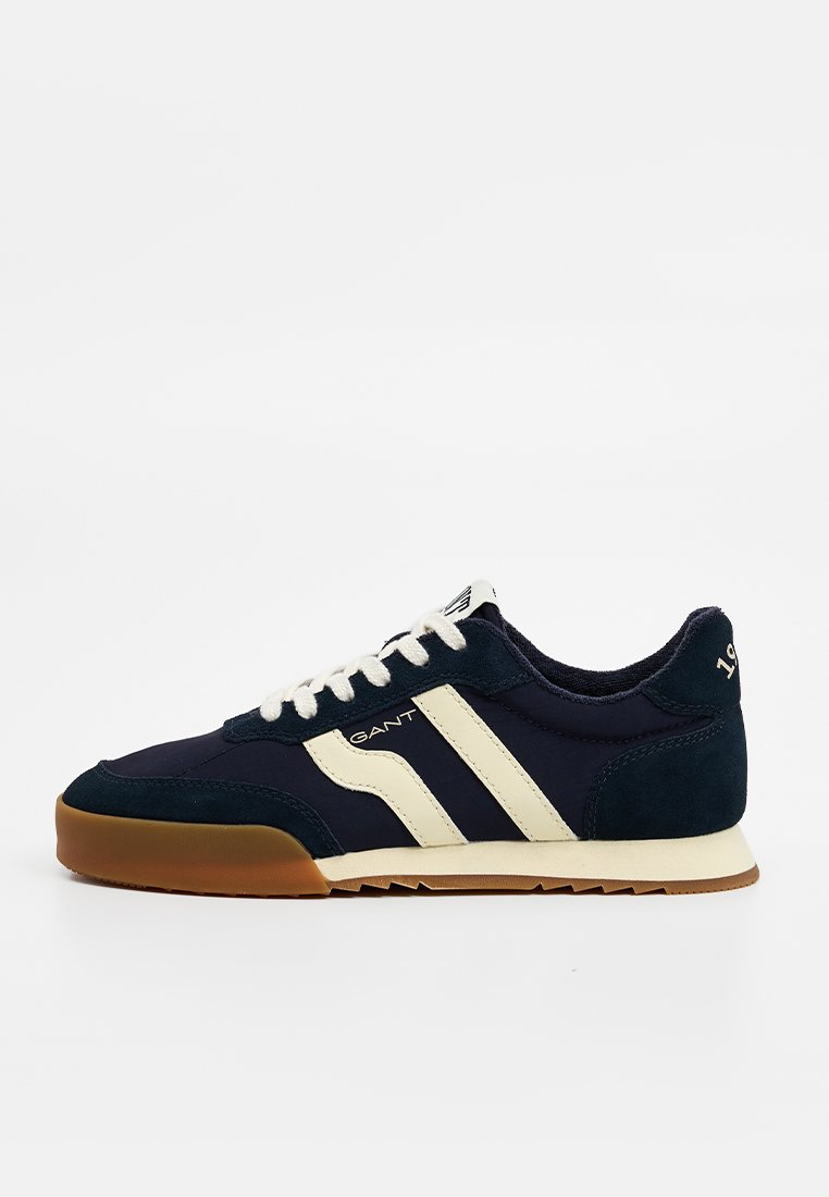 Sneaker blu navy realizzata in suede e tessuto, con strisce crema, lacci bianchi e suola in gomma. Elementi distintivi includono il logo e il dettaglio dell'anno.