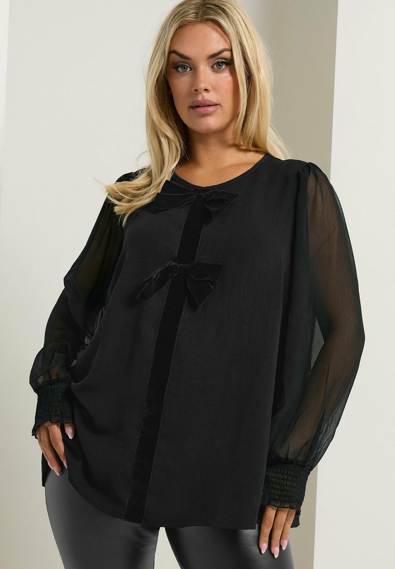 Schwarze Bluse mit transparenten langen Ärmeln, die ein Vorderteil und zwei dekorative Schleifen zeigt. Das Material wirkt leicht und hat eine glatte Textur.
