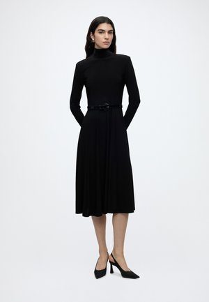 Femme aux longs cheveux foncés portant une robe midi noire à manches longues avec col roulé, une ceinture et des chaussures noires à talons hauts, debout devant un fond blanc.