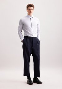Giovane uomo in piedi con le mani in tasca che indossa una camicia da uomo azzurra chiara, pantaloni navy larghi, calzini neri e scarpe eleganti nere.