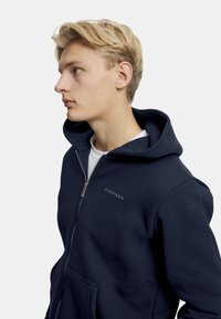 Marinblå zip-up hoodie gjord av mjukt bomullsmaterial, med en kenguruficka, dragsko med huva och tryckt "CASPARA"-logotyp på bröstet.