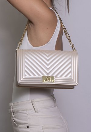 Femme portant un débardeur blanc et un pantalon, portant un sac d'épaule matelassé crème avec une bandoulière chaîne dorée et une fermeture dorée "M MARC ELLIS".