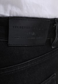 Černé džíny z denimu s texturovaným povrchem a koženou nášivkou s nápisem "SELECTED/HOMME" vyšitým tmavě šedou barvou. Vybaveny standardní zadní kapsou.