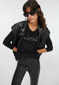 Giacca nera in pelle con spalle a coste, t-shirt nera con pietre decorative e pantaloni neri aderenti. Occhiali da sole neri completano il look.