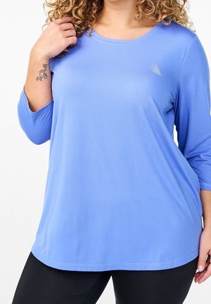Personne aux cheveux bouclés portant une chemise ample à manches longues bleu clair et un pantalon noir, avec un tatouage en écriture sur le poignet gauche.