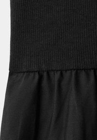 Vêtement noir avec partie supérieure en maille côtelée et tissu tissé froncé en dessous, montrant la texture et le détail des coutures.