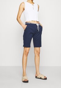 Vit kortärmad linne med vågiga kanter och tre metalldetaljer, kombinerad med marinblå shorts med dragsko och sidofickor.