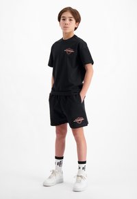 Zwarte katoenen t-shirt en bijpassende shorts, met een grafisch logo in het rood. Witte sneakers met zwart-witte gestreepte sokken. Eenvoudig ontwerp.