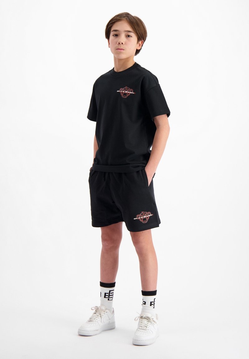 Zwarte katoenen t-shirt en bijpassende shorts, met een grafisch logo in het rood. Witte sneakers met zwart-witte gestreepte sokken. Eenvoudig ontwerp.