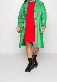 Zelený kožený kabát s černými knoflíky, přes červené šaty. Černé šněrovací boty a malá kulatá zelená ta ška s texturovaným designem. Kárované akcenty.