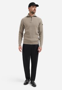 Lichtbruine geribbelde pullover met een ritskraag, gecombineerd met zwarte broeken en zwarte schoenen. Pet op het hoofd, handen aan de zijkant.