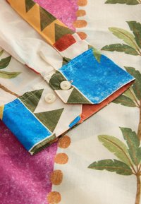 Poignet de chemise à manches longues avec motifs géométriques et feuilles tropicales en bleu, vert, orange et rose, avec deux boutons blancs.