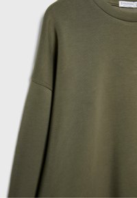 T-shirt à manches longues vert olive avec col rond, couture d’épaule visible et étiquette de la marque Stradivarius à l’intérieur du col.