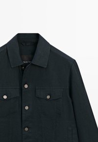 Veste noire à boutons avec col et deux poches poitrine à rabat avec boutons métalliques, présentée sur fond blanc.