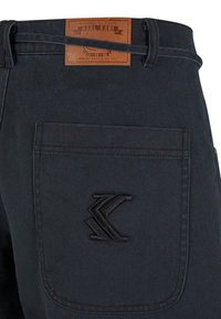 Dark denim pants featuring a leather label with embossed text, back pocket with a black embroidered logo, and a classic waistband design.